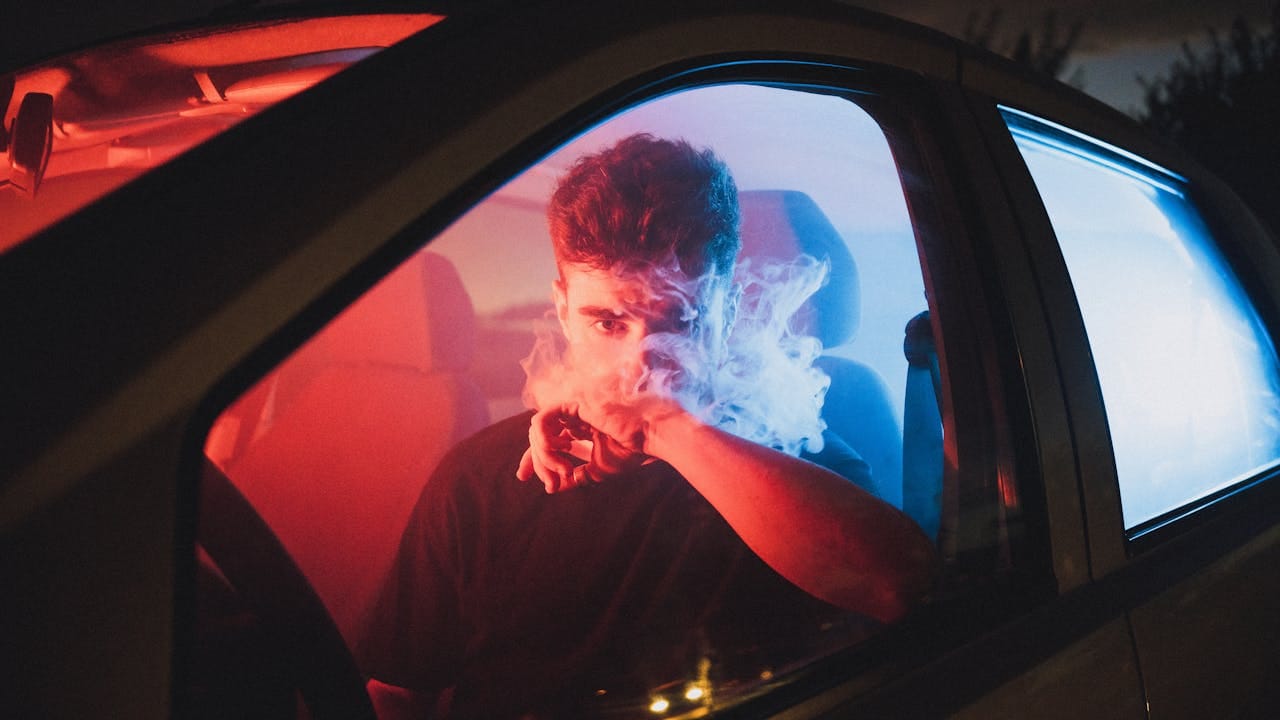 my-steps Moody image of a young man vaping inside a car with vibrant red and blue lighting.